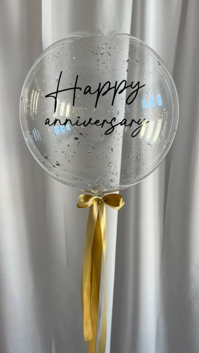 Globo burbuja transparente con mensaje de aniversario, confeti y un lazo dorado.